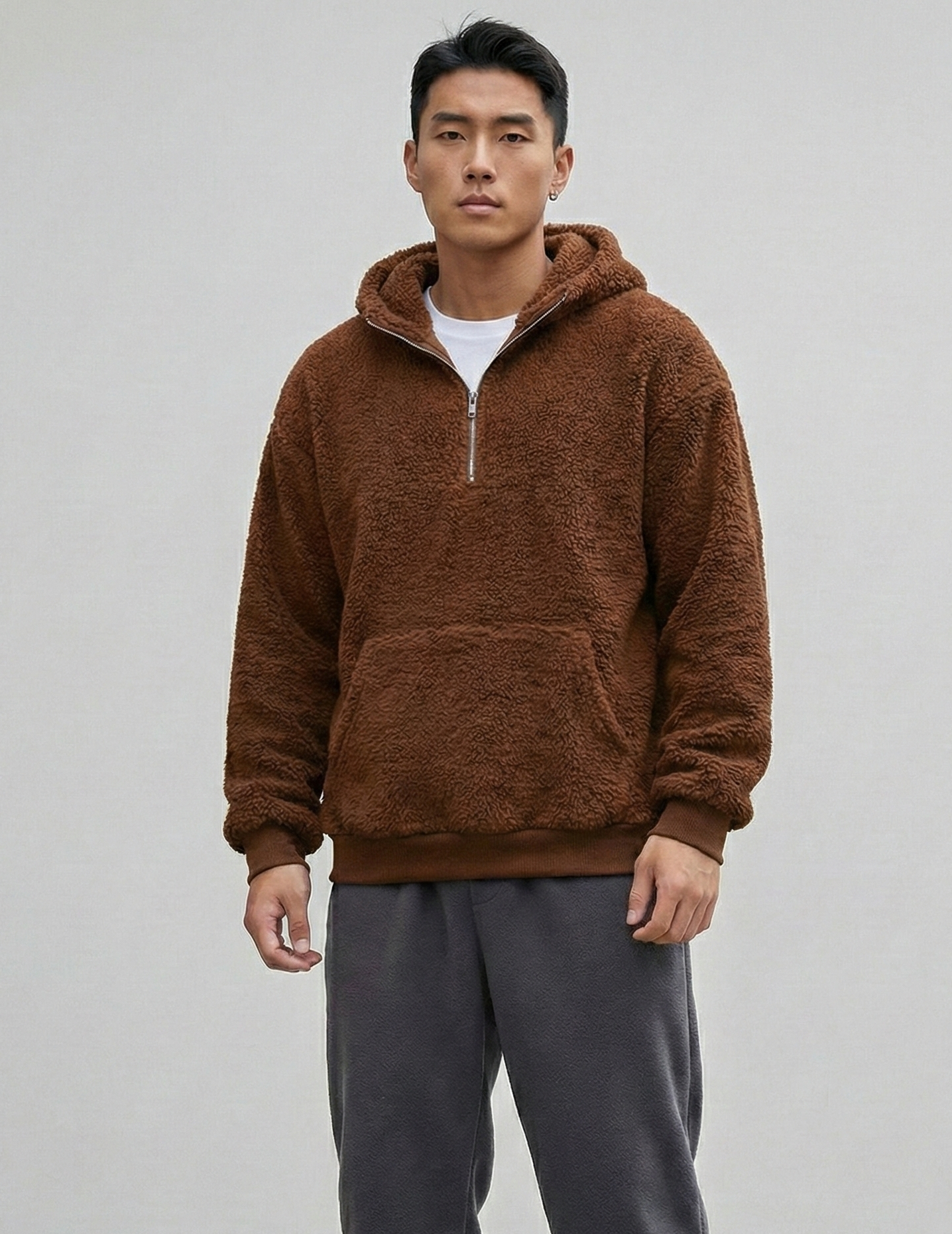 Man wearing a brown fleece pullover and gray sweatpants with blue sneakers on a plain background